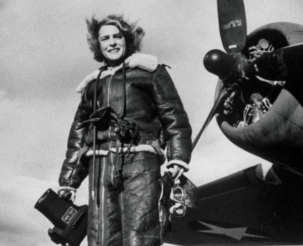 Black and white photo showing a woman in a pilot's jacket with a camera, standing in front of airplane propeller